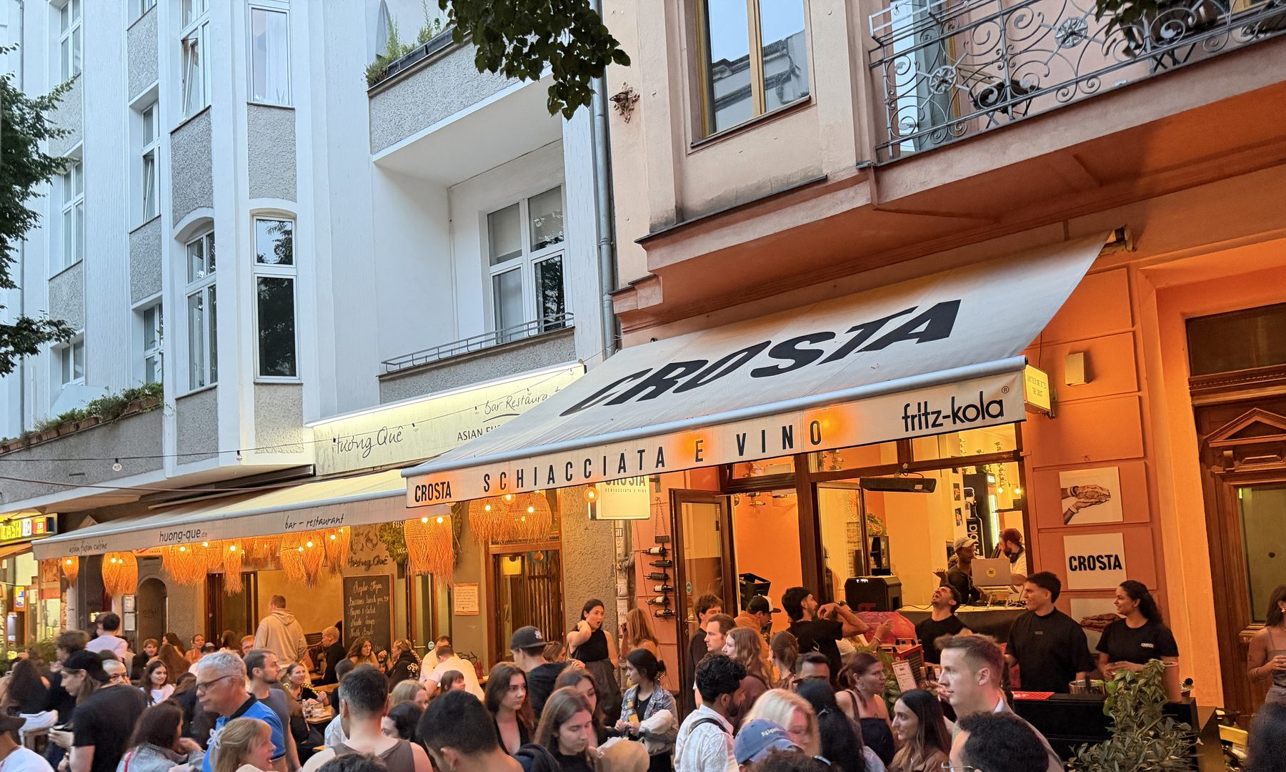 A packed summer evening outside Crosta on Weinbergsweg, terrace full and the awning lit up