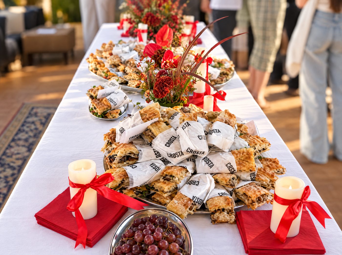 Long catering table in Berlin with platters of Crosta schiacciata bites wrapped in Crosta paper, red flowers and candles